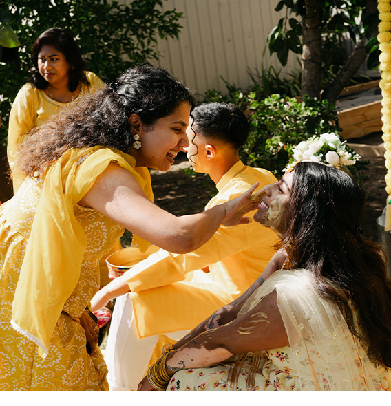 Indian family in yellow attire smile spreading paste on face of bride at wedding ceremony.