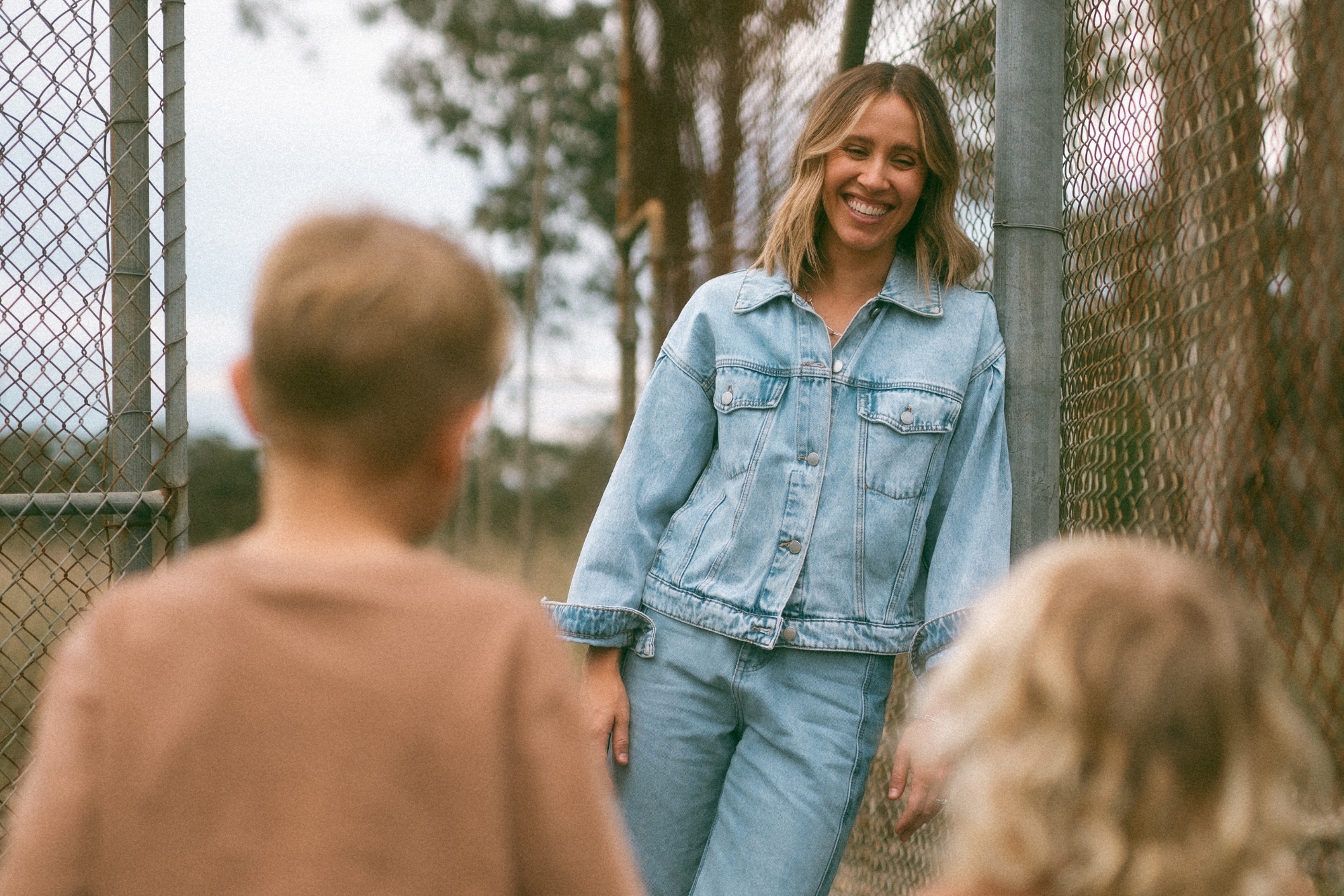 Mother leans on chain fence smiling looking at her sons.