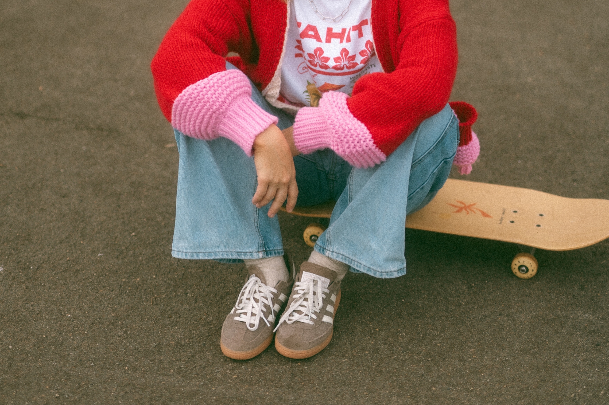 Woman in bright colored streetwear sits on skateboard on concrete court.