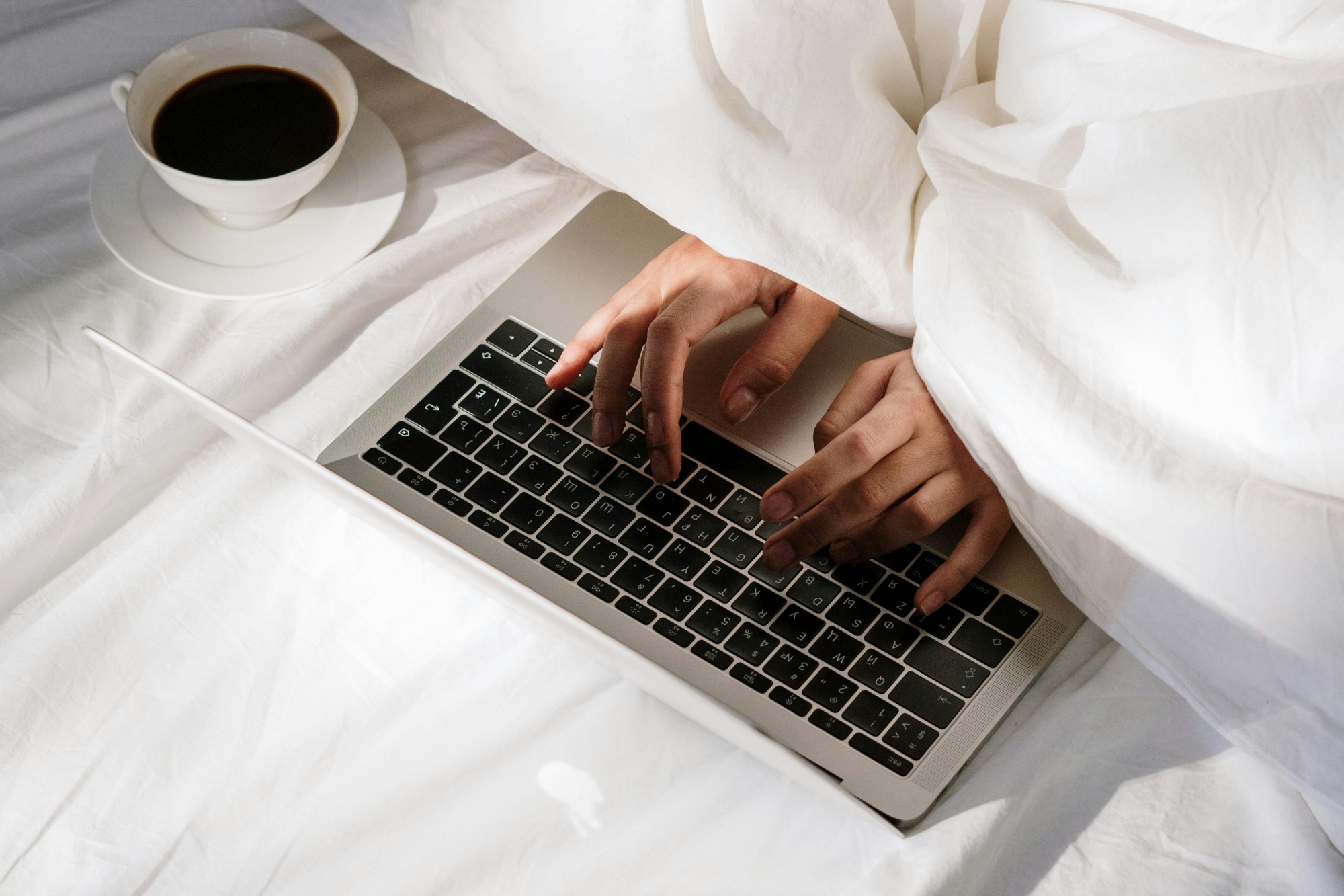 Typing on Mac laptop while covered in white linen bedsheets under dappled lighting, with coffee sitting next to laptop.