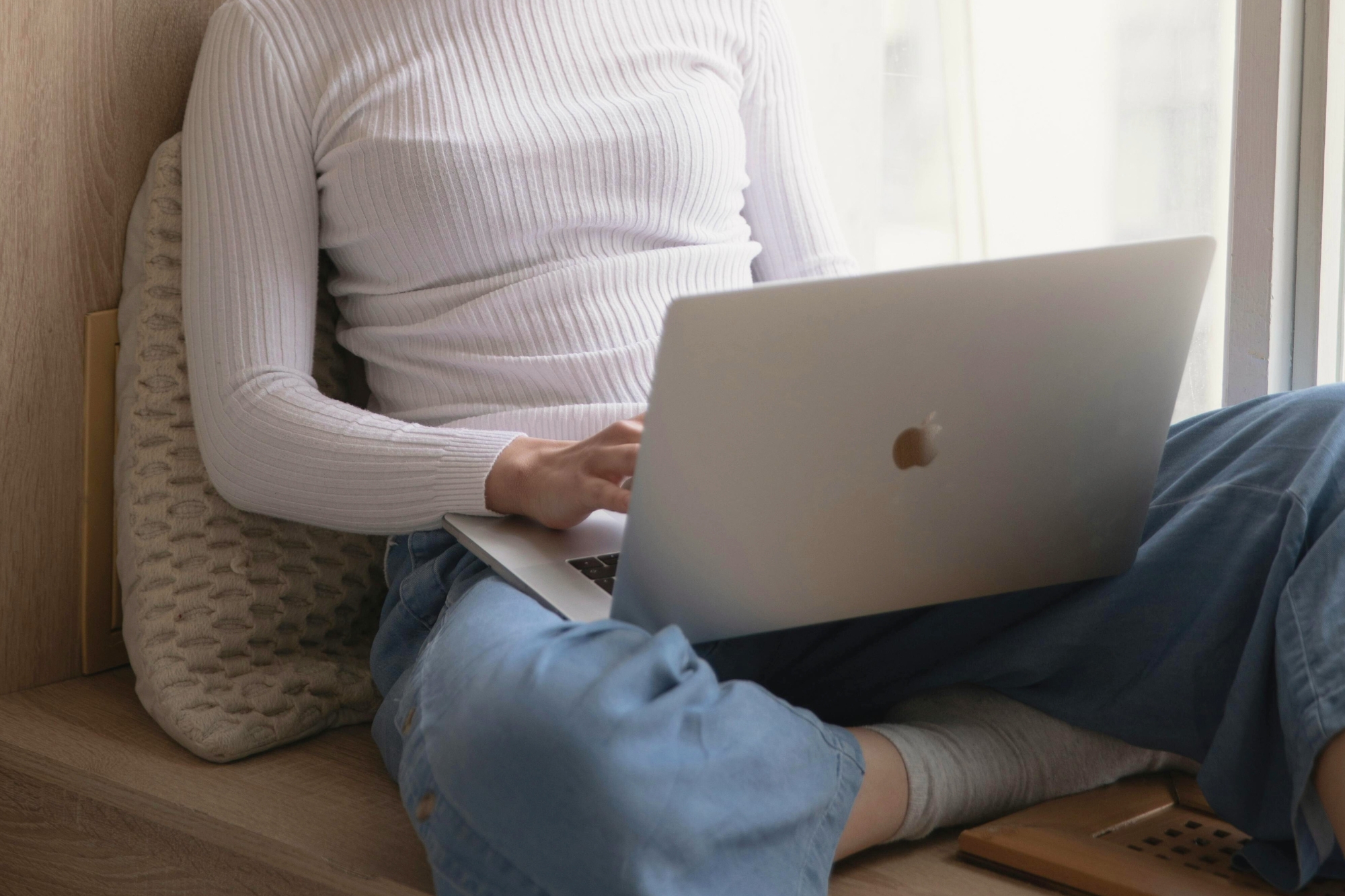 Woman sits on edge of window sill typing on Mac laptop.