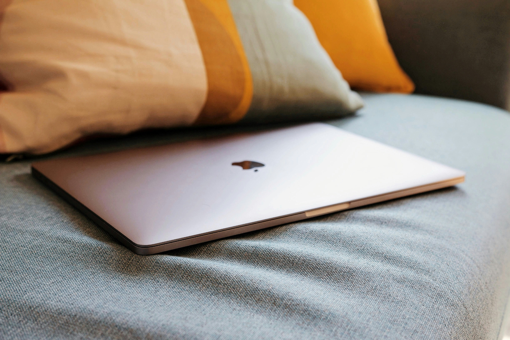 Rose gold Mac laptop closed laying on grey couch with mustard hued pillows.