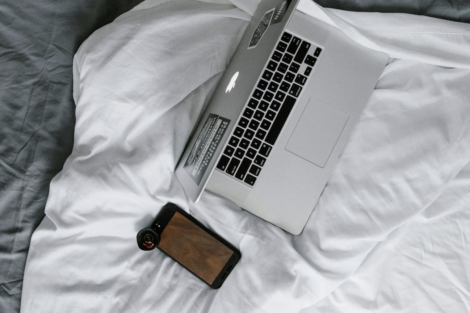 Mac laptop covered in stickers next to iPhone with camera lens attached, both sitting on white and grey bed sheets.