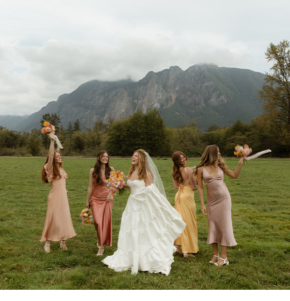 Bride smiling with bridesmaids wearing blush toned dresses standing in a field under overcast skies.