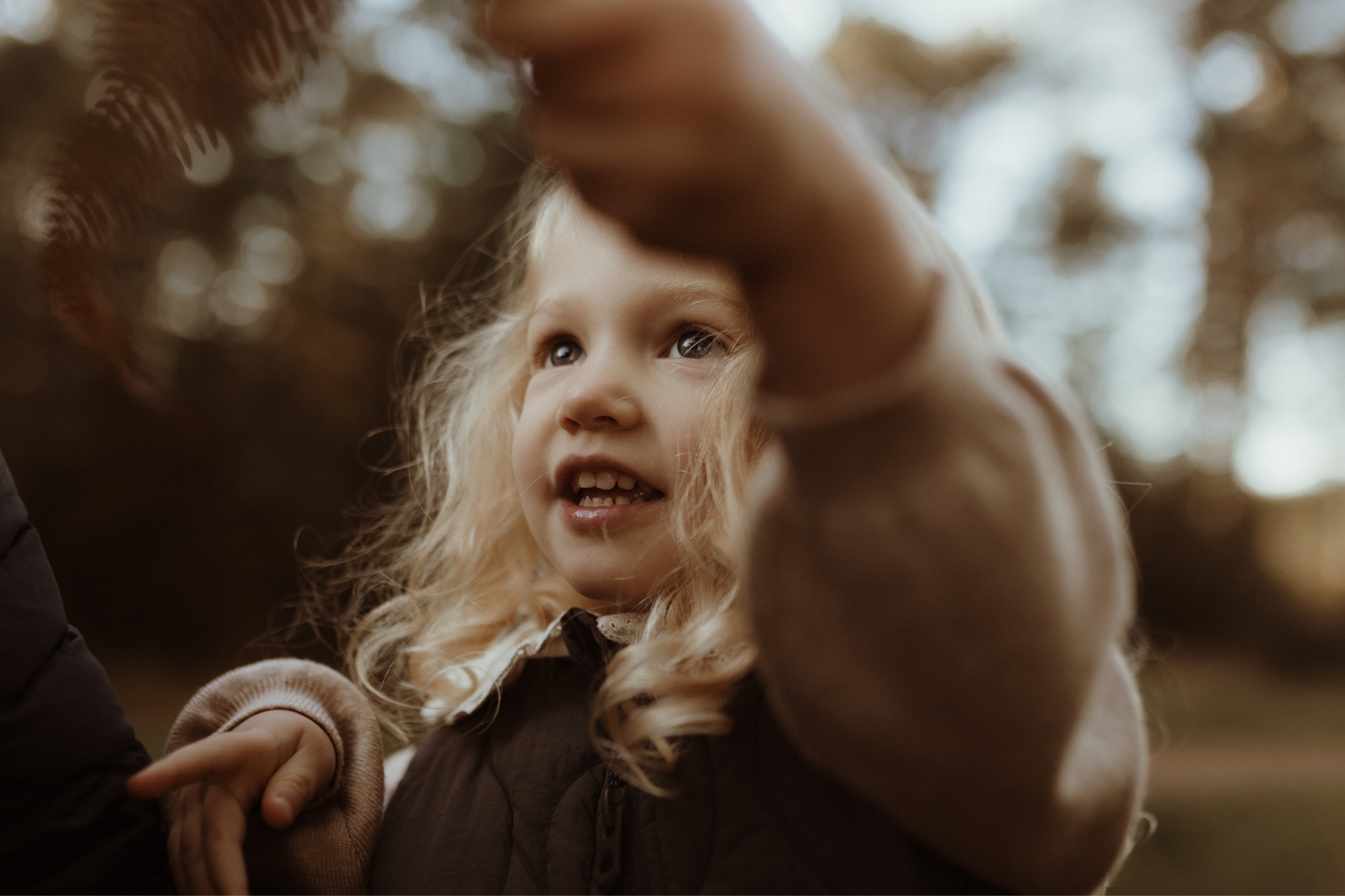 Close up of young blonde girl reaching for trees with blurry background.