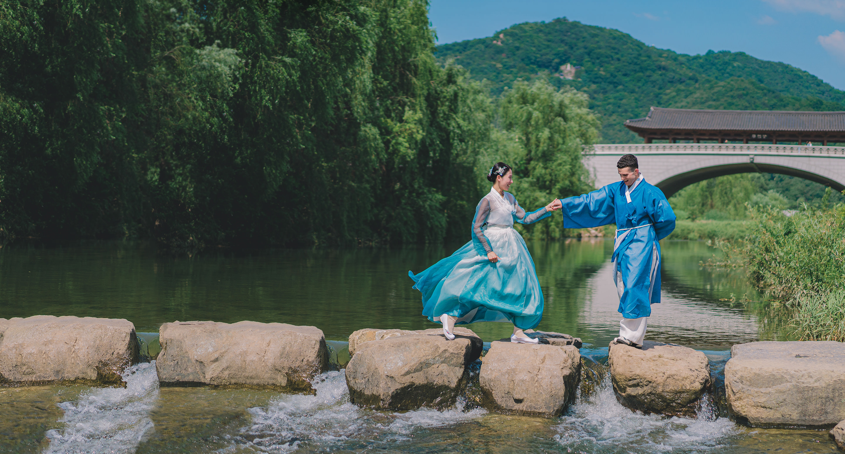 Couple coréen en tenue de mariage bleue, se tenant par la main, traversant un pont de pierres au-dessus d’une rivière.