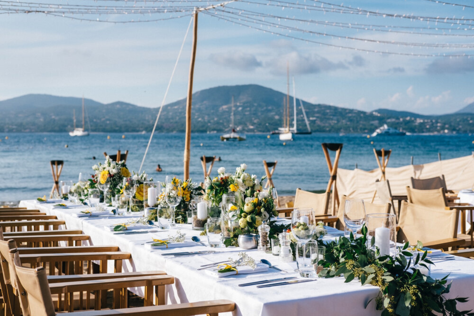 Outdoor wedding dining and seating with white and yellow flower centerpieces overlooking sailing harbor under sunlight.