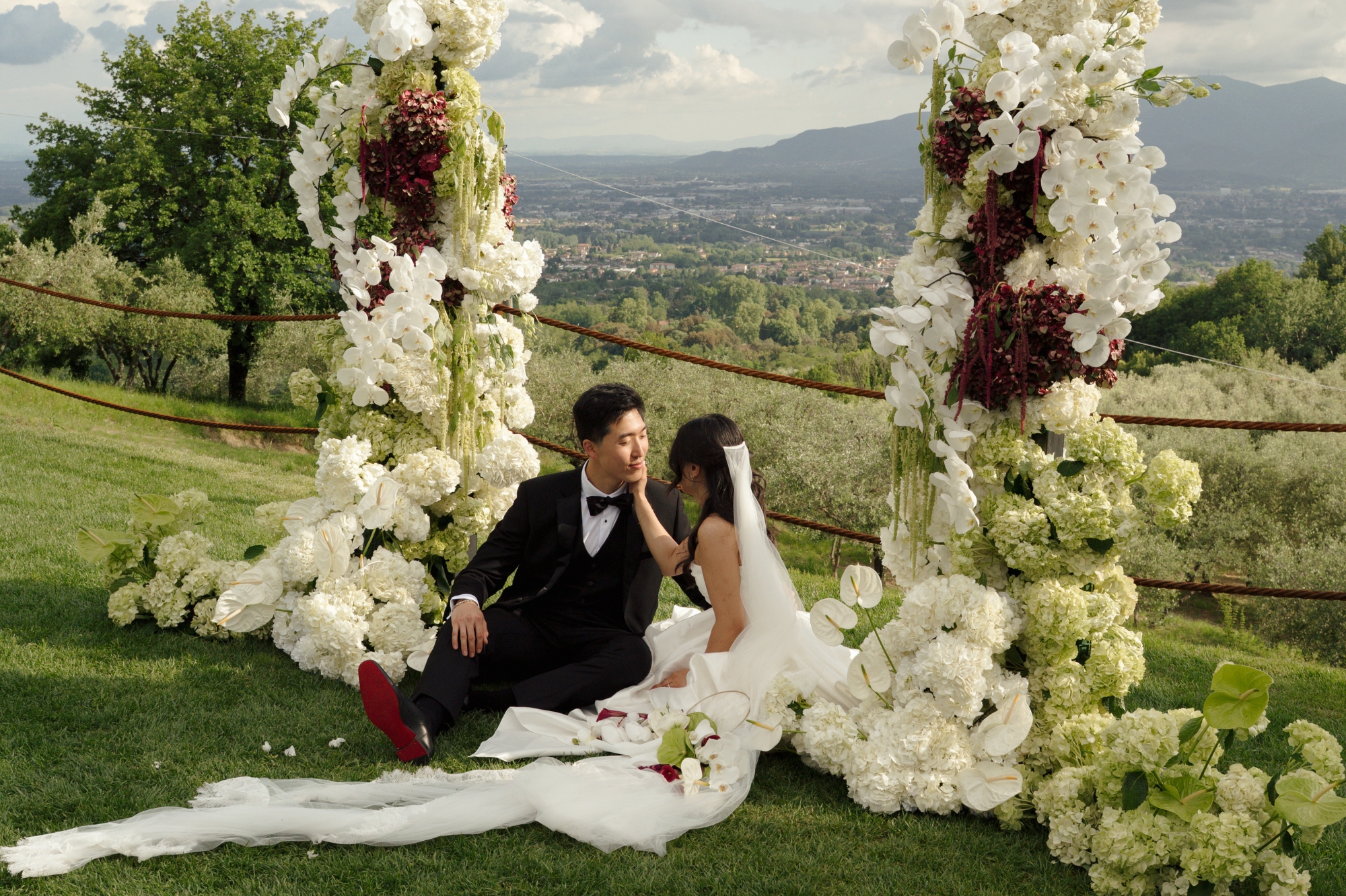 A newlywed couple seated on grass gaze at each other under wedding arch covered in white chrysanthemum flowers.