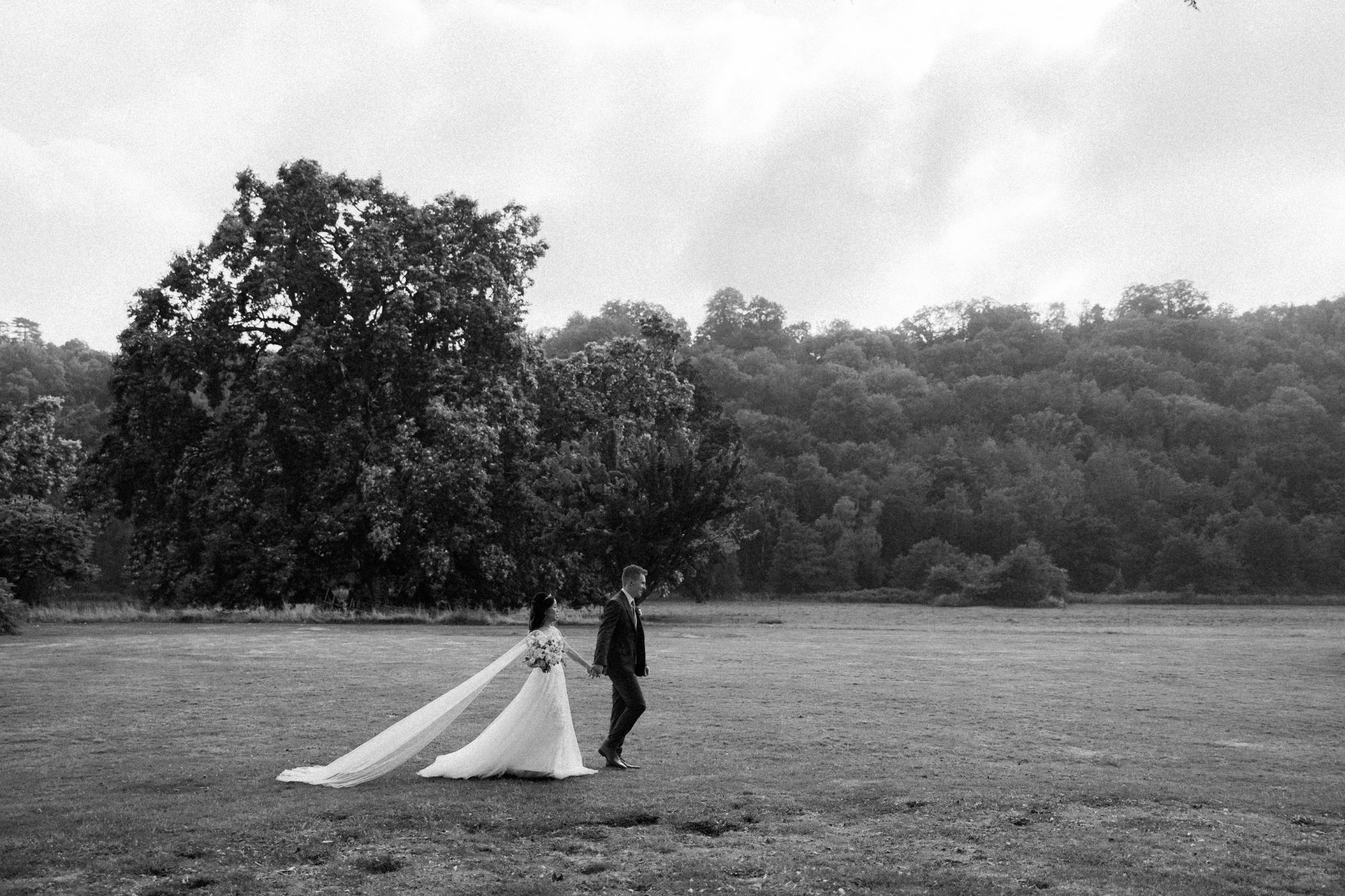 Bride and groom hold hands walking across wide grassy park field together.