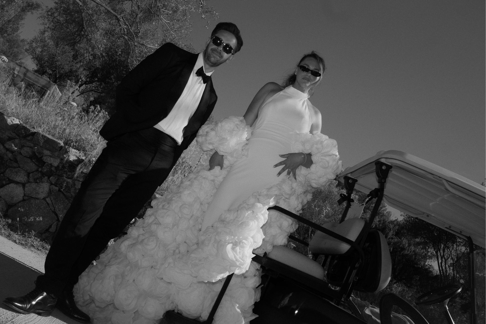 Modern newlywed couple pose with serious expressions wearing sleek, sophisticated wedding attire and sunglasses standing by golf cart.