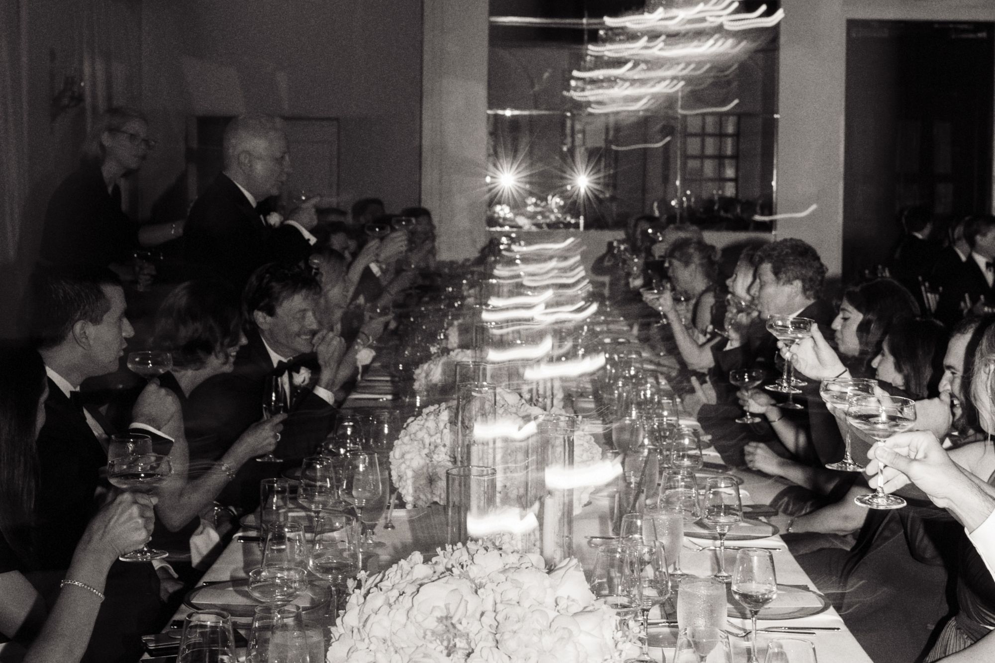 Black and white photo of wedding guests at long dining table with candlelight at reception.