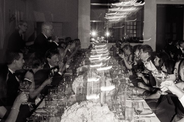 Black and white photo of wedding guests at long dining table with candlelight at reception.