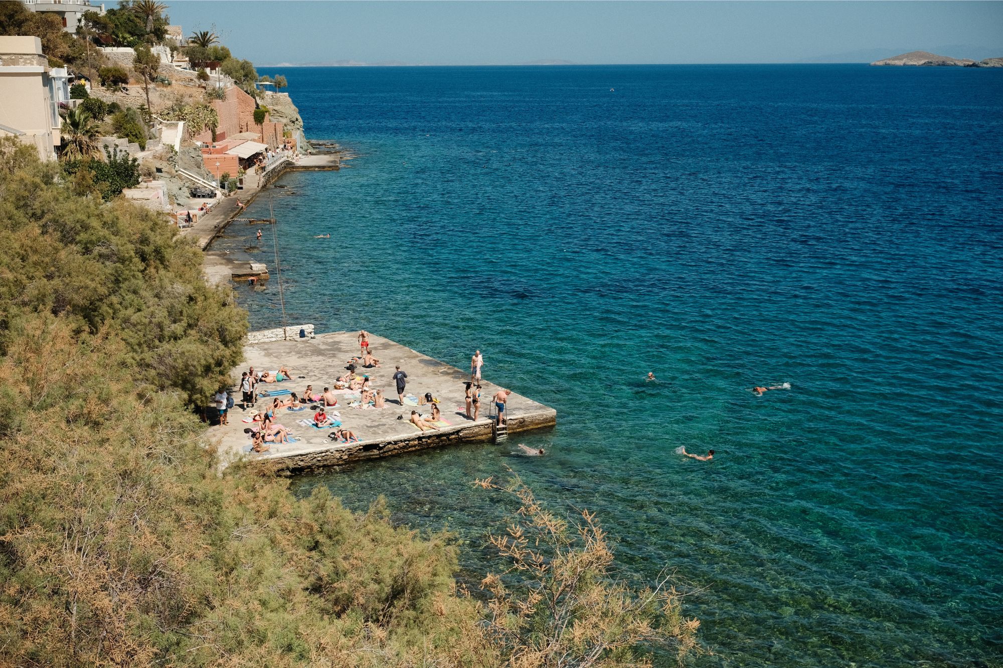 Sunbathers in Italy lie under bright sunlight on concrete pier, some jumping in water and paddling around.