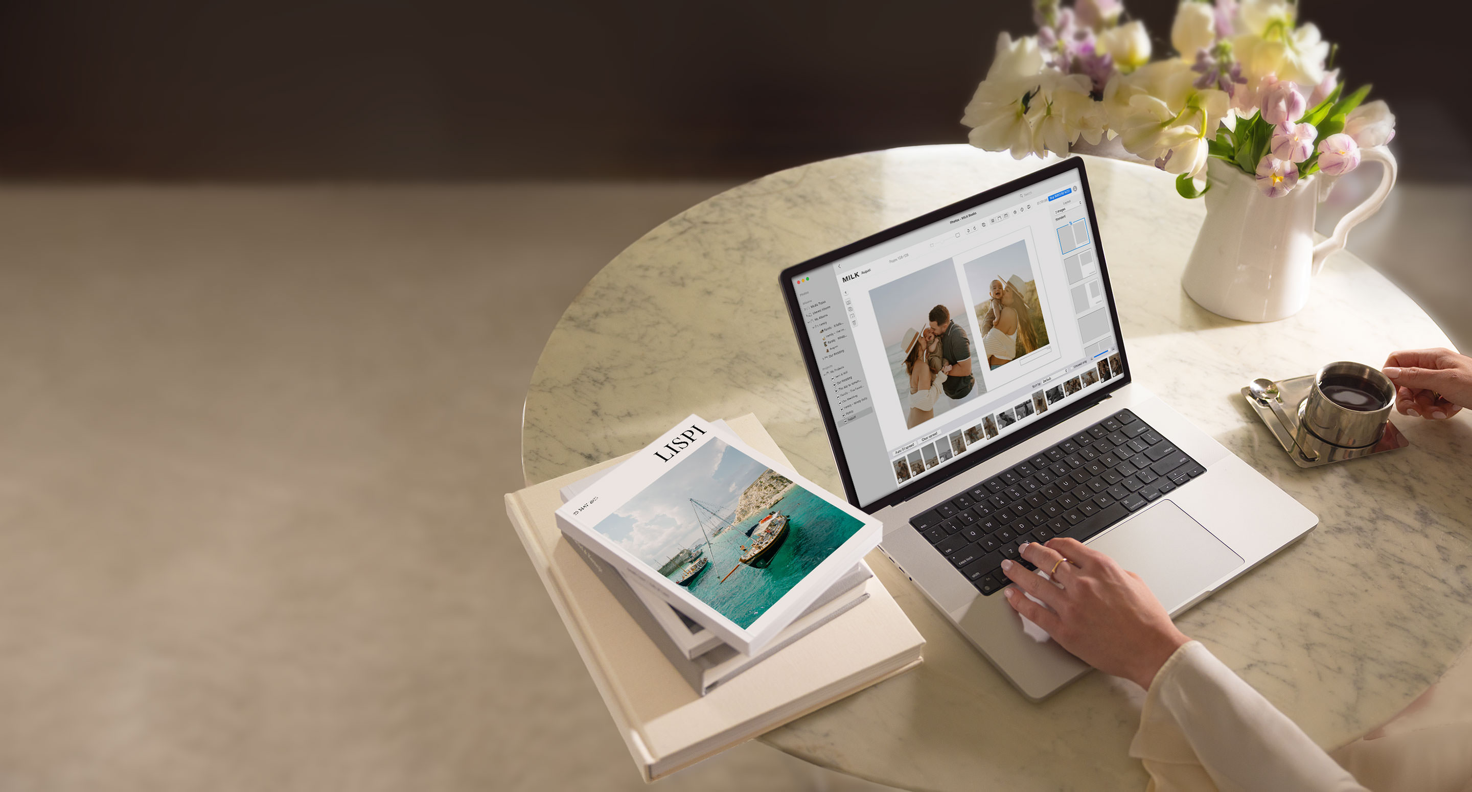 Eine Person gestaltet ein Fotobuch von MILK Books auf ihrem MacBook, mit Büchern und einer Tasse Kaffee auf einem Marmortisch.