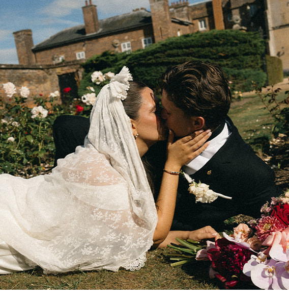 Newlywed couple kiss amongst flowers from bouquet while lying on grass.