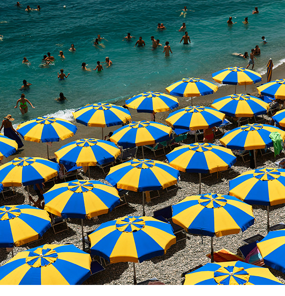 Bright blue and yellow sun umbrellas cover stony beach with many swimmers in shore water.