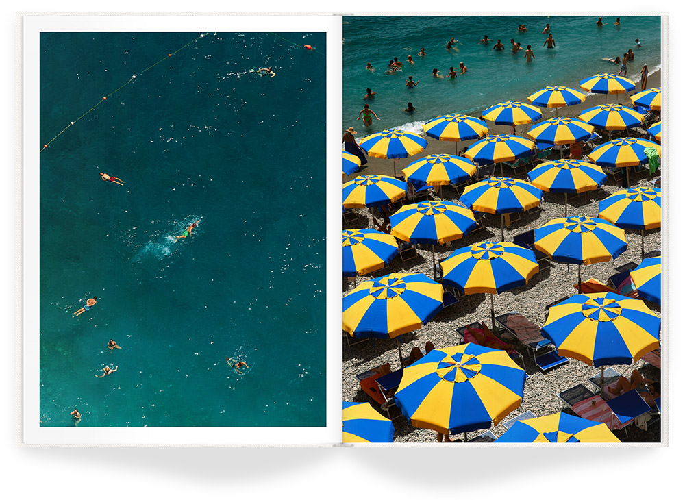 Bright blue and yellow sun umbrellas cover stony beach with many swimmers in shore water.