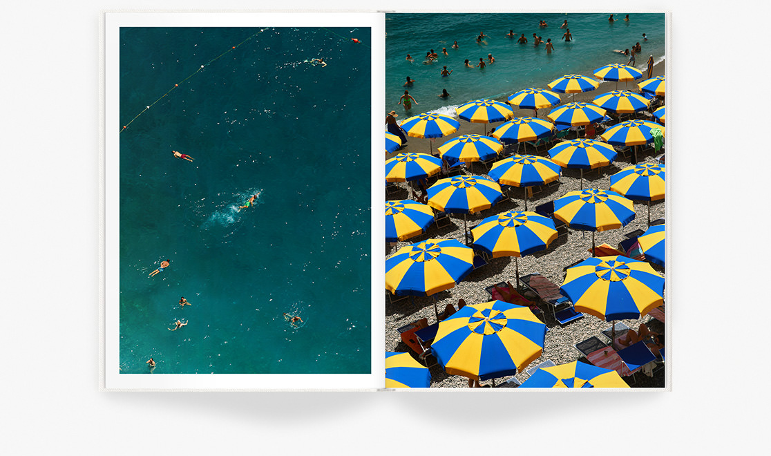 Bright blue and yellow sun umbrellas cover stony beach with many swimmers in shore water.