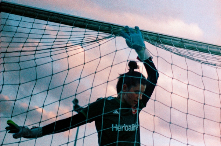 Female soccer player goalie pushes hands against football goal net, exhausted under sunset behind cloudy sky.