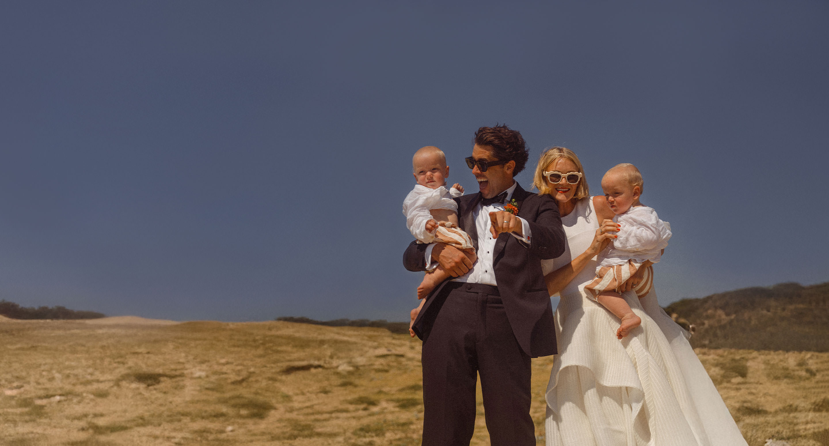 Pareja de recién casados sonriendo juntos en una colina mientras sostienen a dos bebés con atuendos blancos.