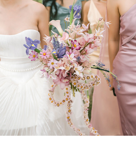 Bride standing next to bridesmaid in pink holding colorful pastel flower bouquet in shades of pink and lilac.