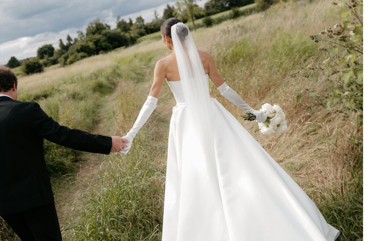 Bride walks hand in hand with groom across grass paddock in bridal attire holding white flower bouquet.