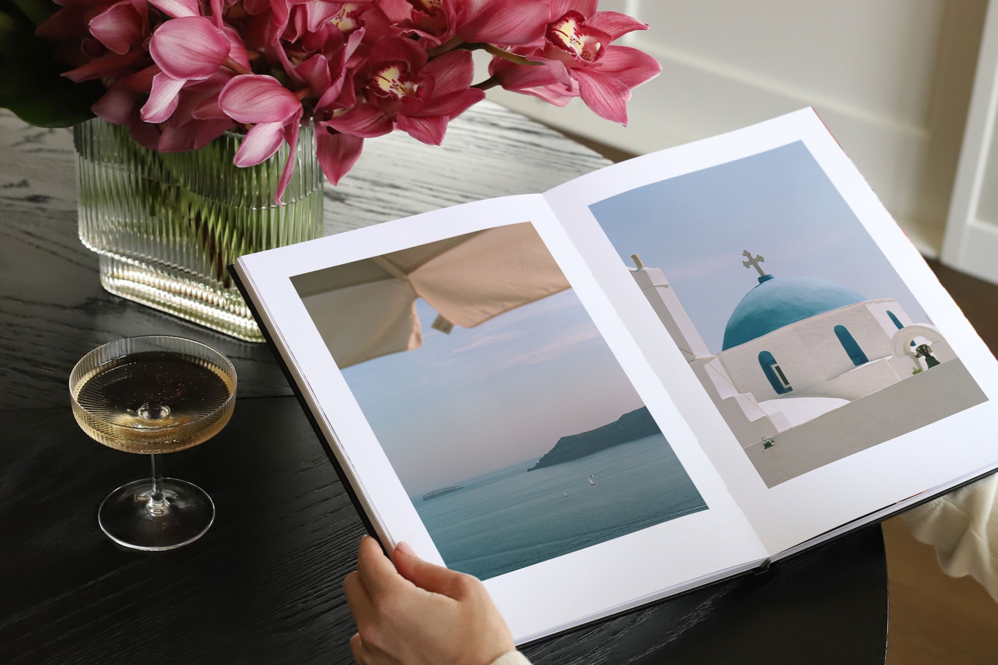 Woman holds a large Premium Photo Book with travel images inside, champagne in a glass sitting on table next to the book.