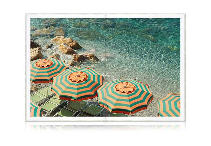 Open Portrait Premium Photo Book showing bright beach with clear turquoise water and matching green and orange umbrellas on the shore.