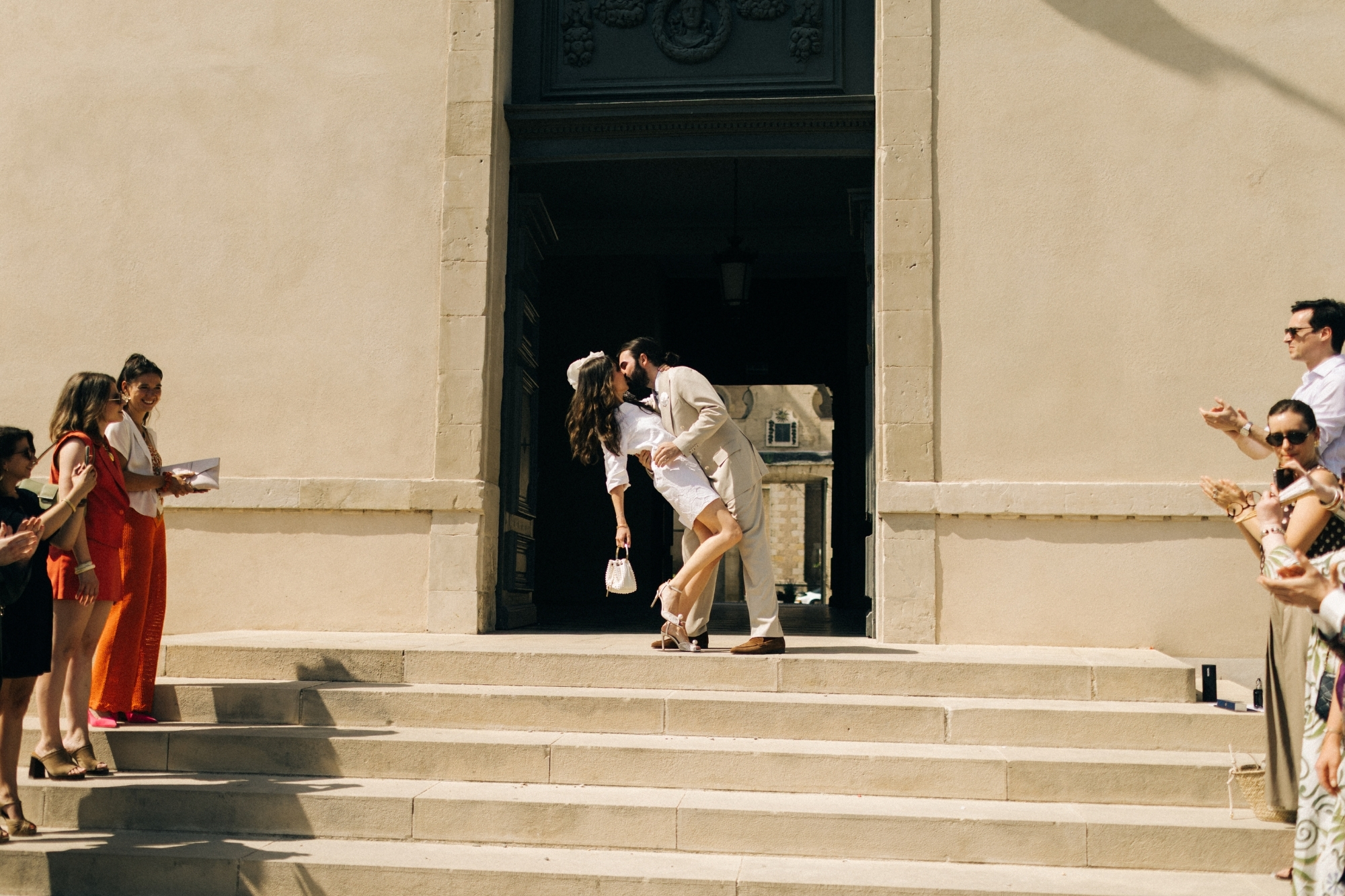Newlywed couple kiss outside of office building with loved ones clapping lining the stairs.