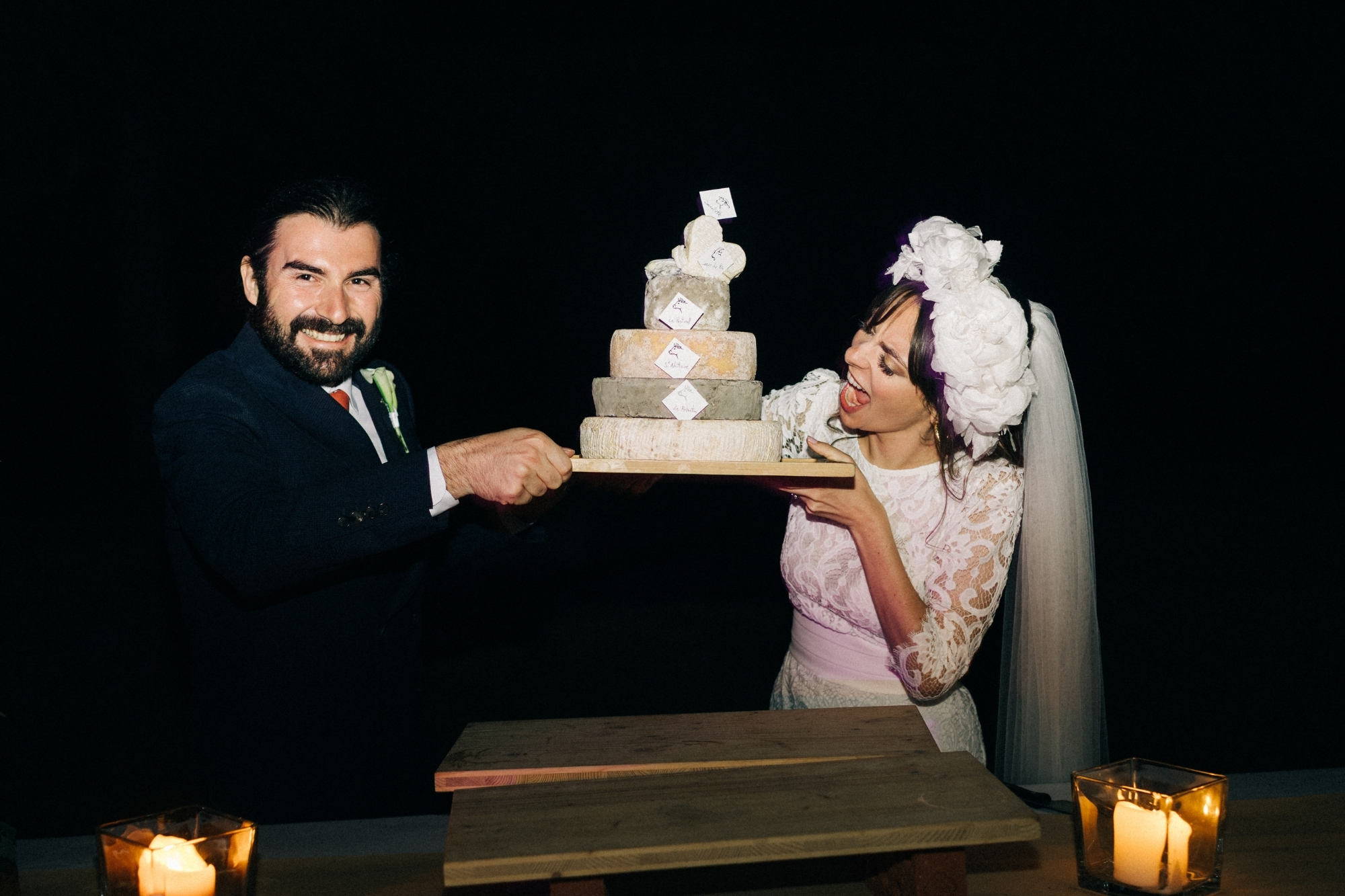 Newlywed couple holding cake made up of 5 tiers of different cheese wheels.