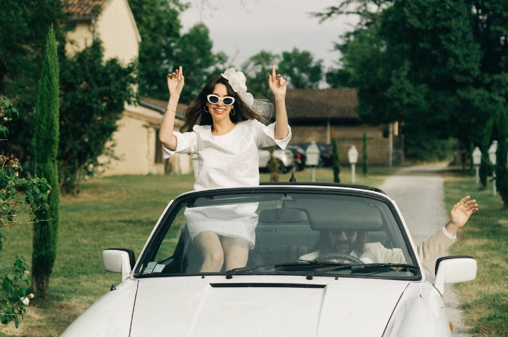 Bride wearing white sunglasses sits outside of white convertible car and waves her hands as groom drives.