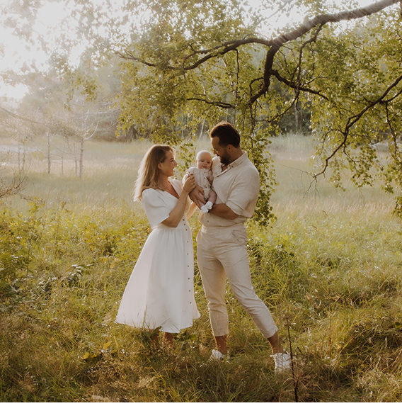 Parents wearing white standing in overgrown misty grass field in bright sunlight, holding a baby together.