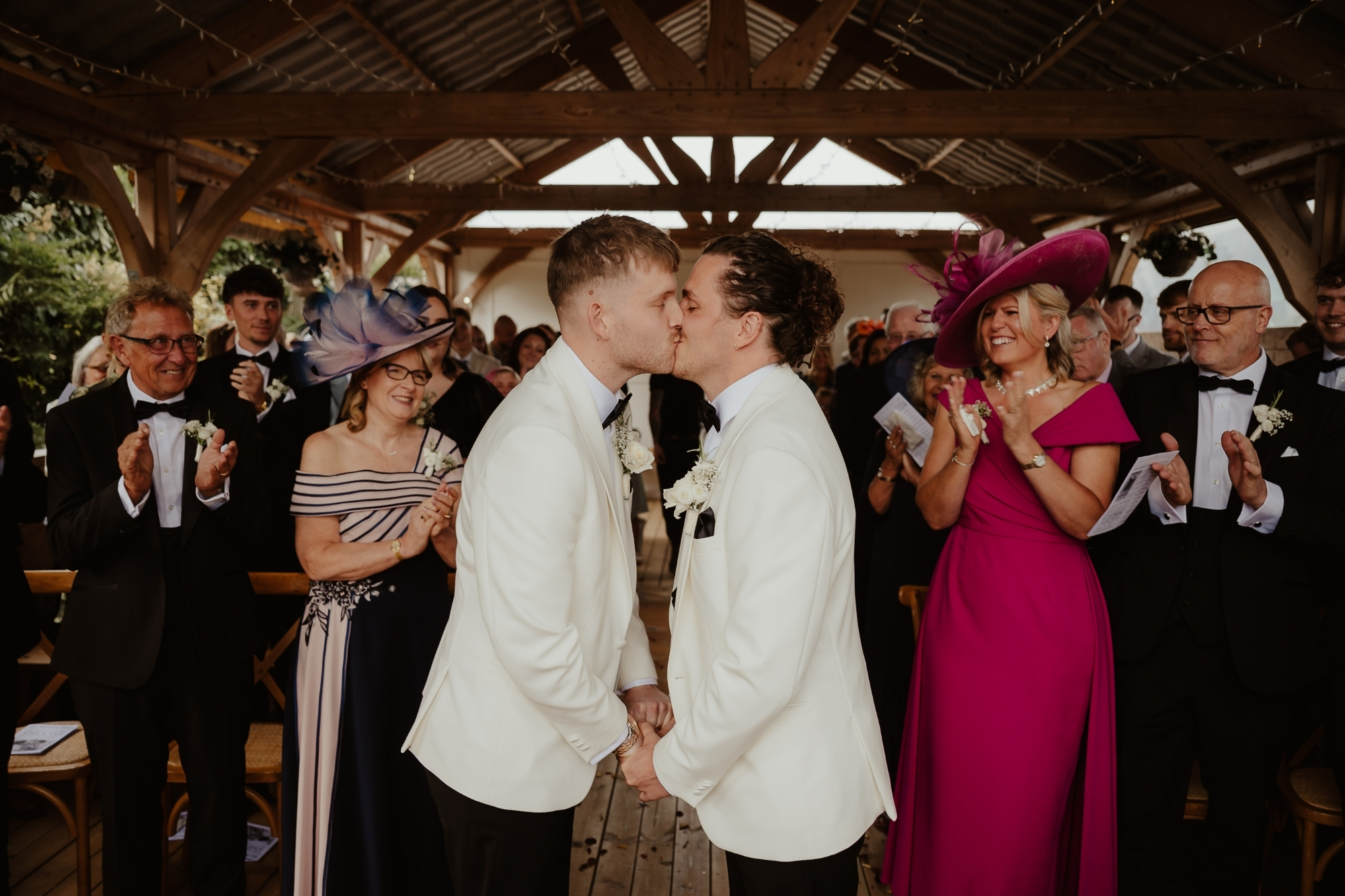 Two grooms kiss at end of aisle while loved ones smile on.