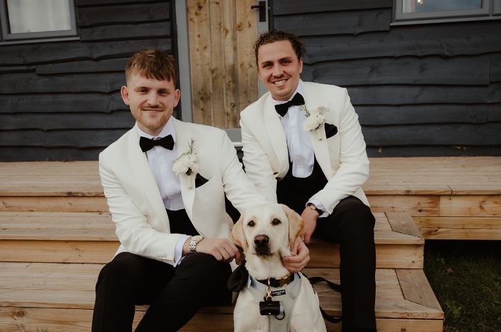Two grooms sit together wearing white suit jackets with a dog sitting in front of them.
