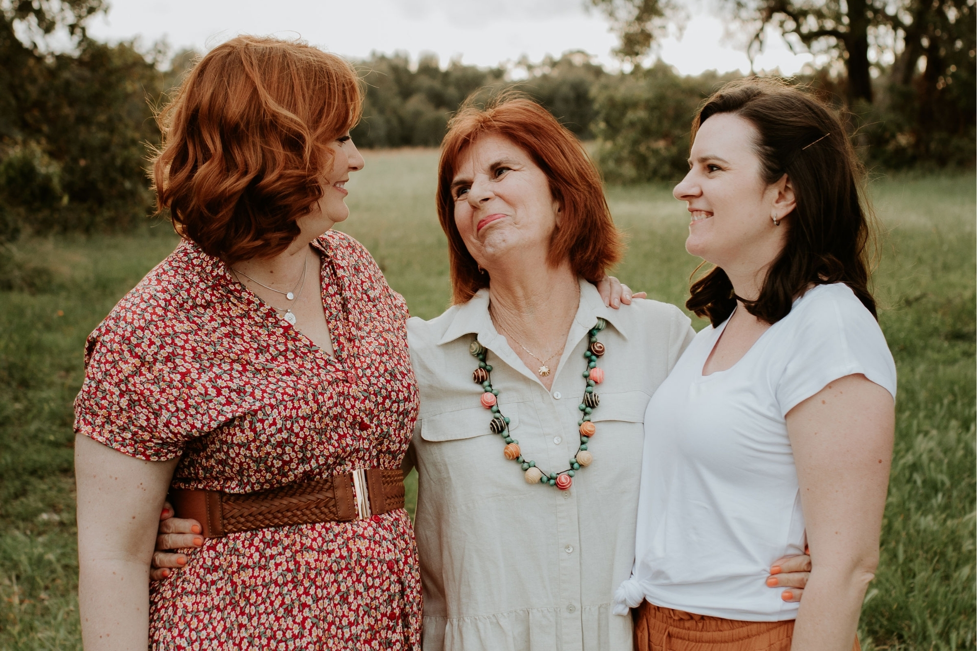 Three women of different generations in the family smile and hold each other while looking at each other.