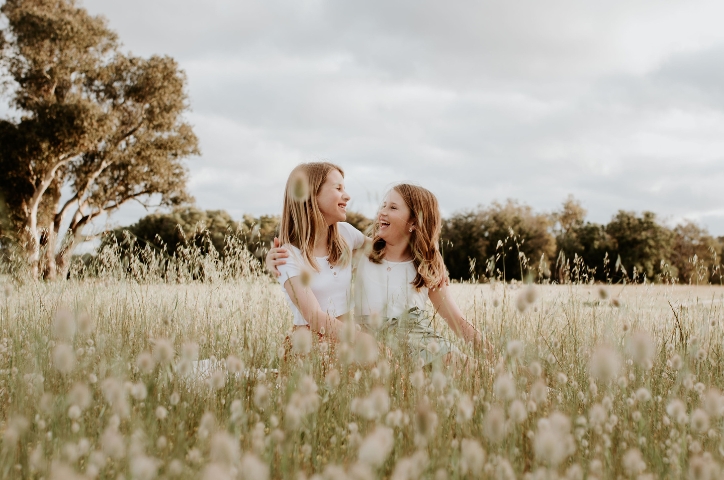 Two girls smiling at each other sitting in cotton field.