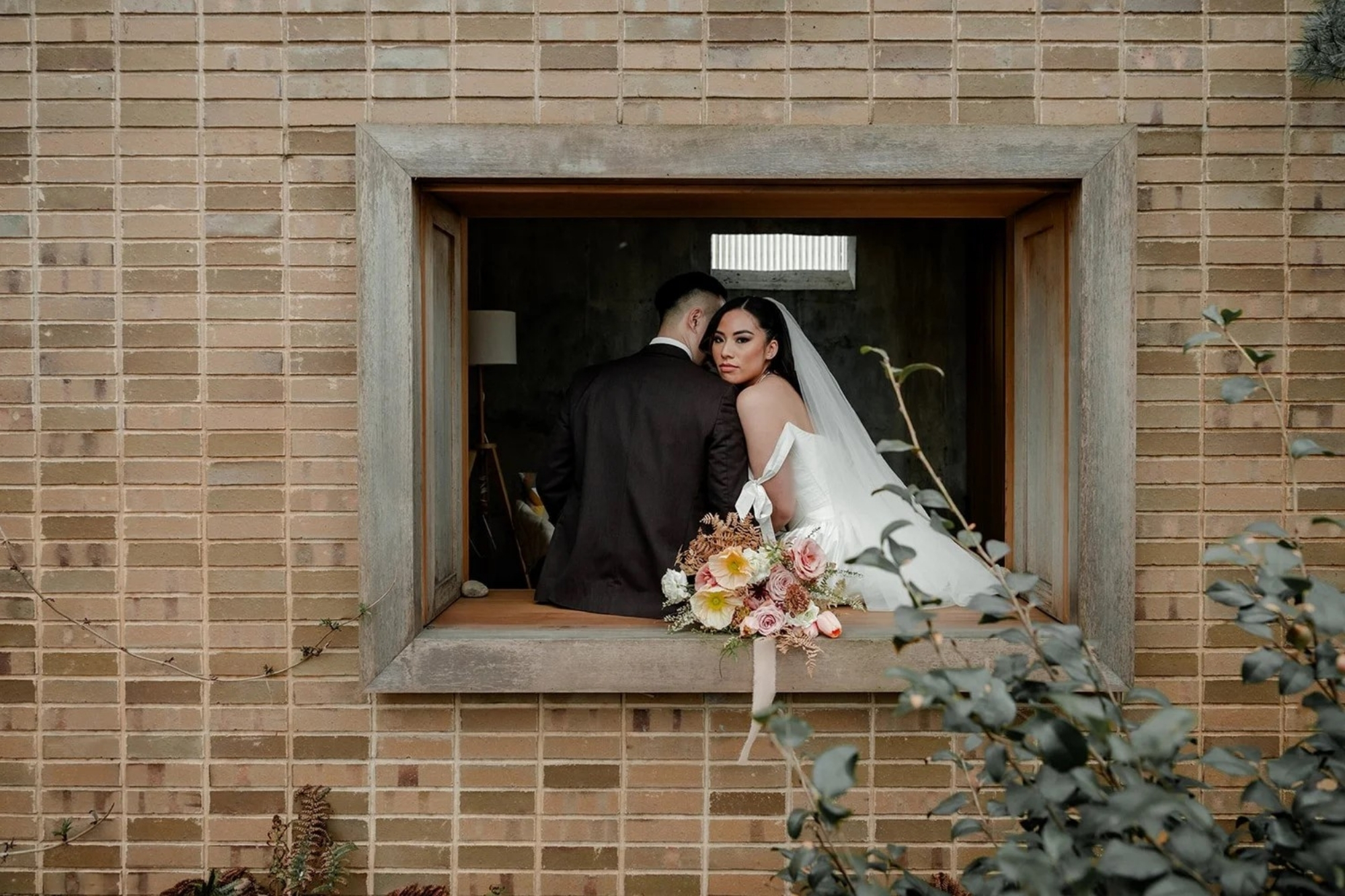 Newlywed couple sitting together in windowsill built into brick wall.