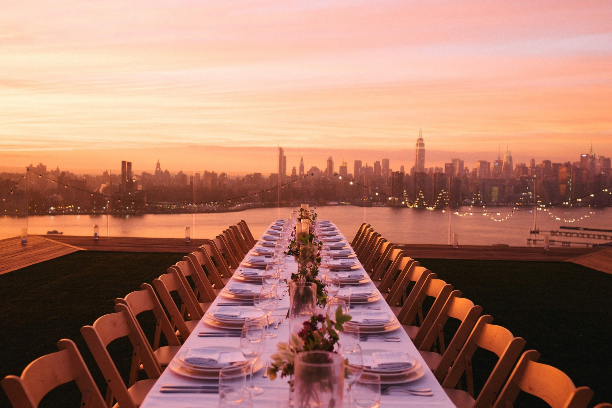 View of New York beyond a dining table set on a rooftop under a pink and orange sunset.