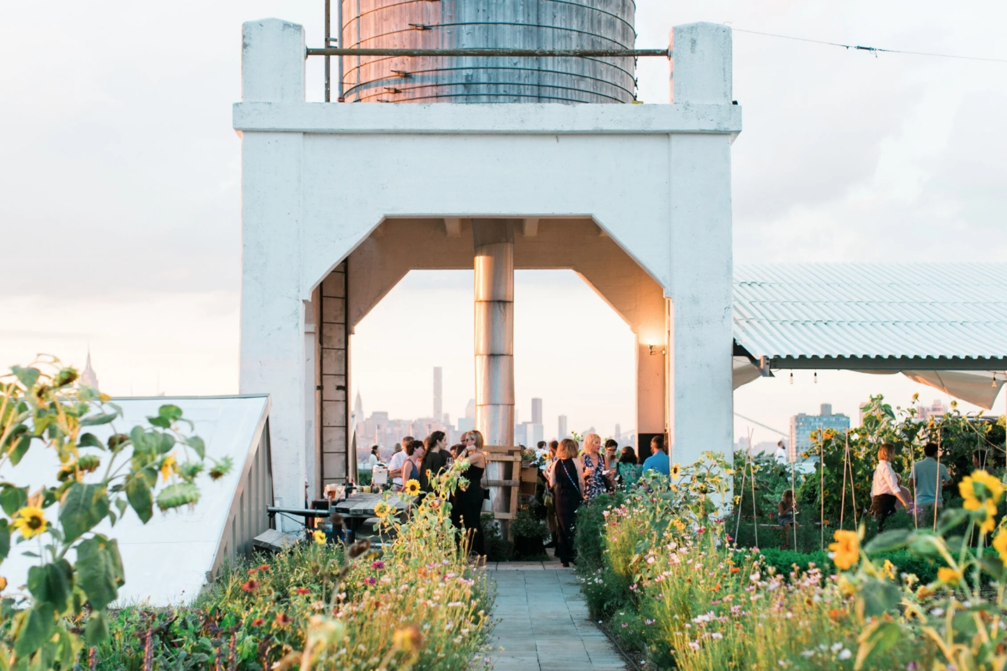 Guests dine and socialize amongst garden of flowers under shed with view of New York City on horizon.
