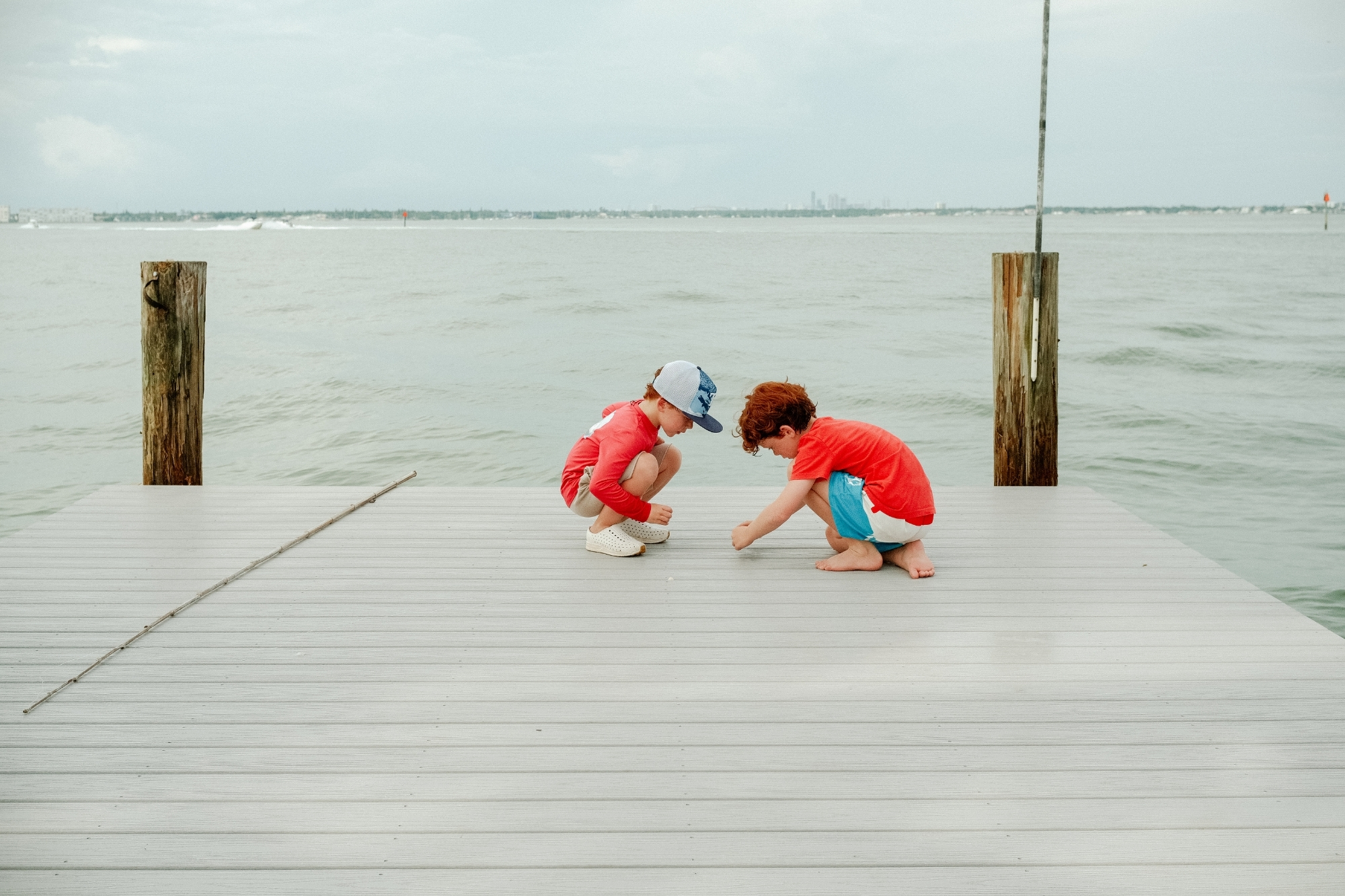Two boys squatting preparing fishing line at end of pier on cloudy day.