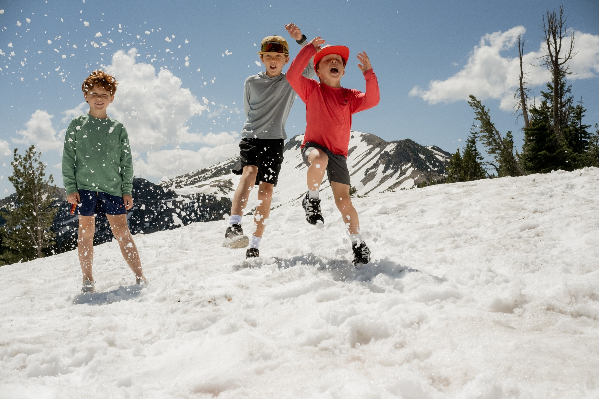 Three boys smiling and skipping through heavy snow while under bright sunlight on a mountainside.