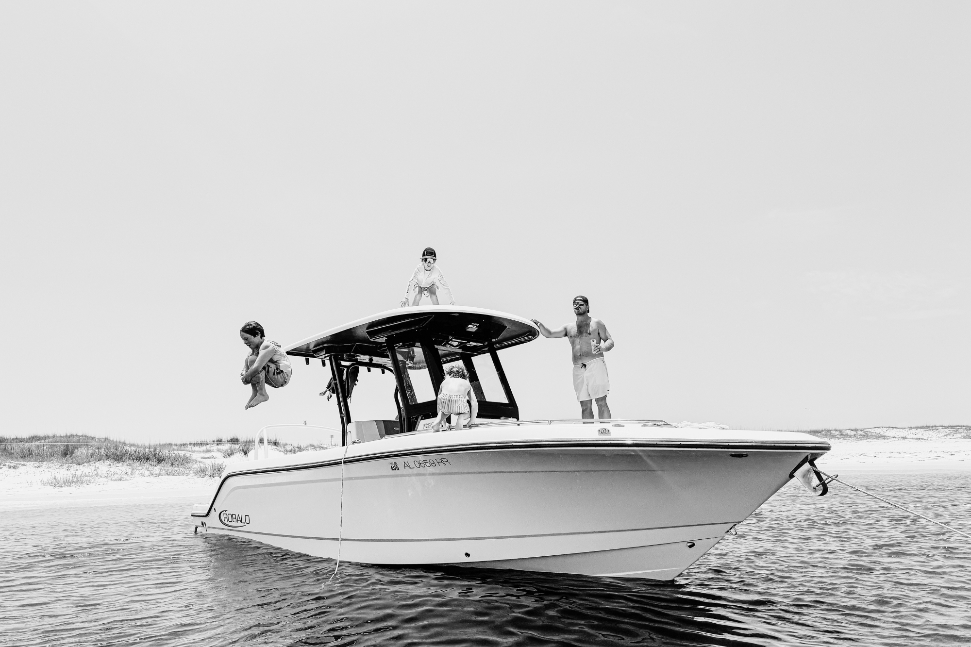 Kids jumping off a boat into ocean near a short, with dad holding a drink and watching over them.