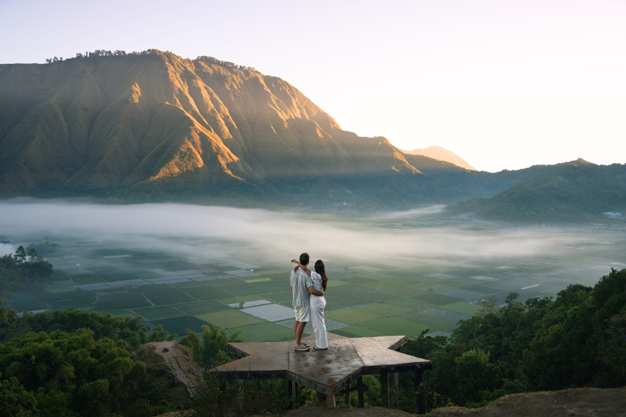 Couple standing on platform holding each other while looking over viewpoint of hilly valley at sunset.