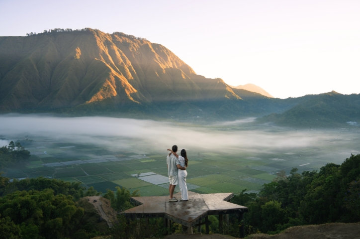 Couple standing on platform holding each other while looking over viewpoint of hilly valley at sunset.