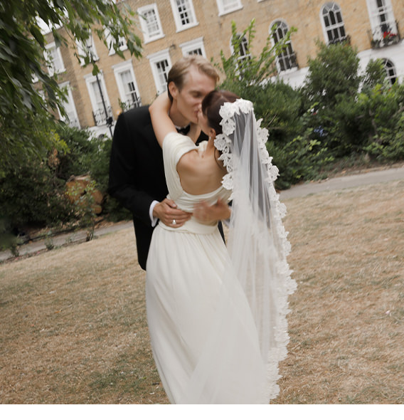 Newlywed couple in wedding attire kiss in the middle of a public park field.